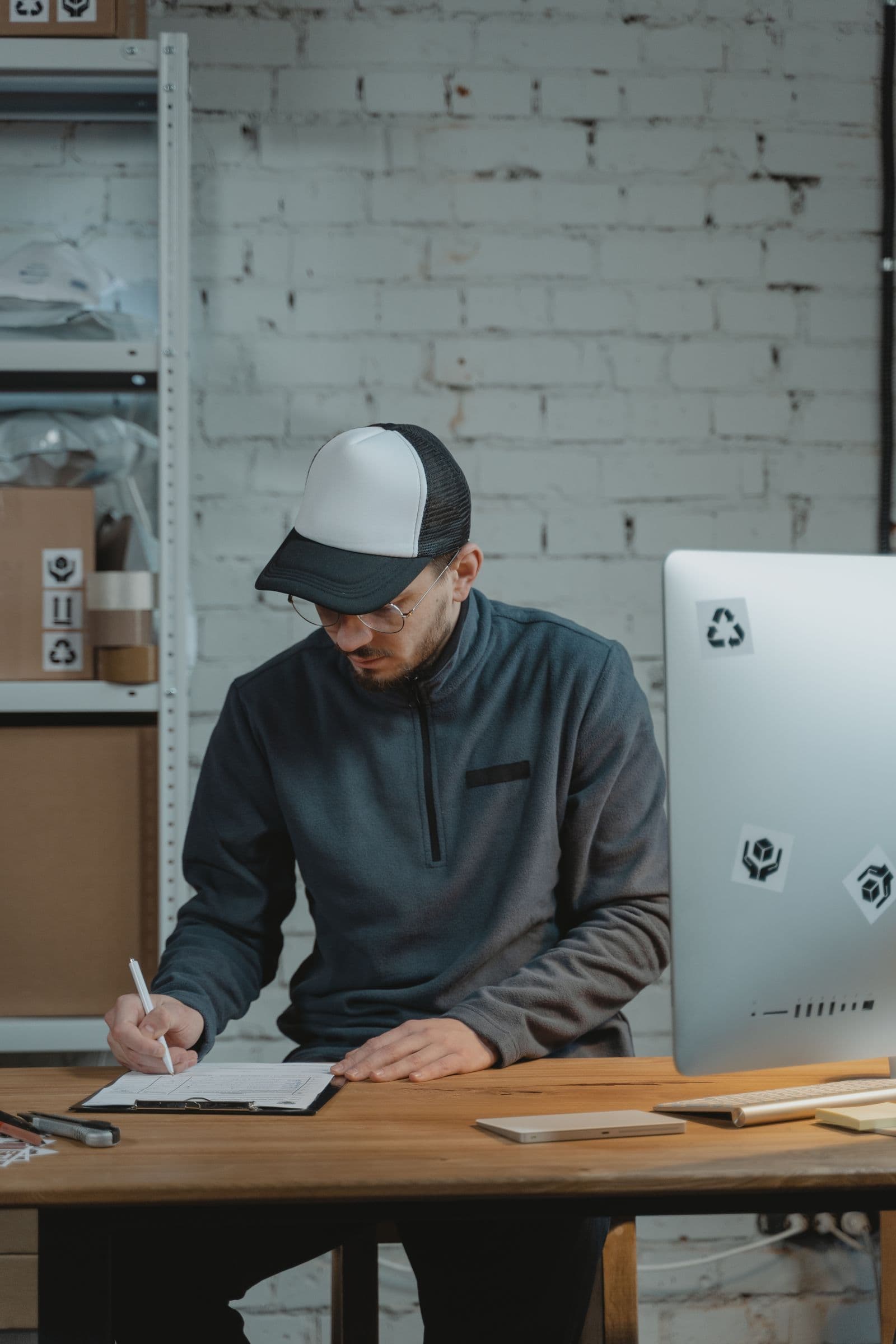 A logistics worker completing a paper form on a clipboard beside a desktop computer