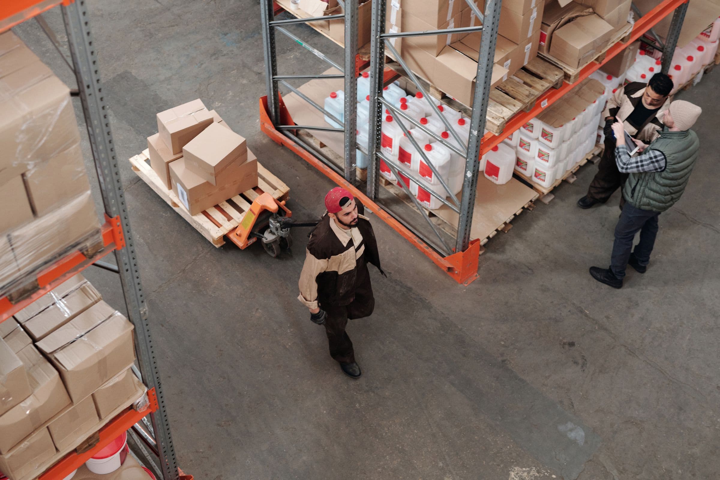 Overhead view of a working warehouse floor: racking, pallets, a worker with a pallet jack and a supervisor holding a tablet